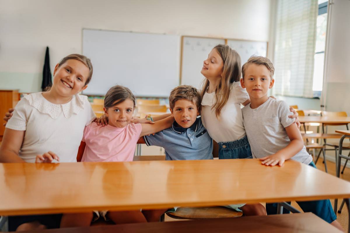 Five smiling elementary age children hugging and posing together in a bright classroom, depicting friendship and learning